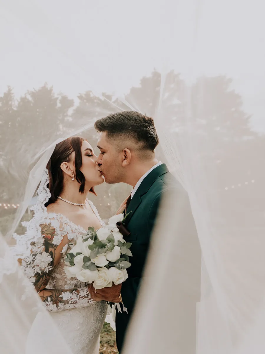Beso de los novios bajo el velo, en el jardín de Laureal con la neblina de la tarde